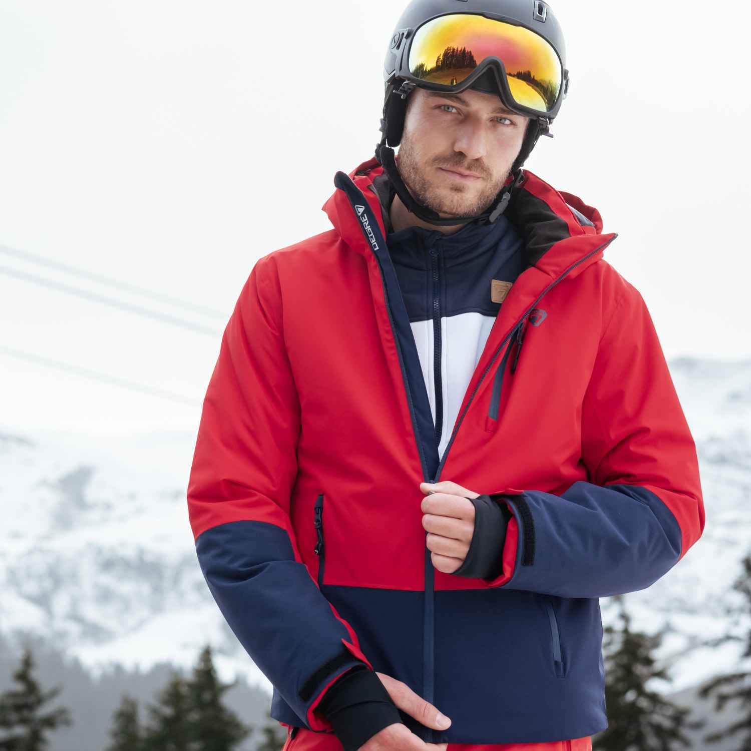 A man wearing ski clothing standing in the snow. He's zipping up a ski jacket to stay warm, wearing ski goggles in the cold weather.